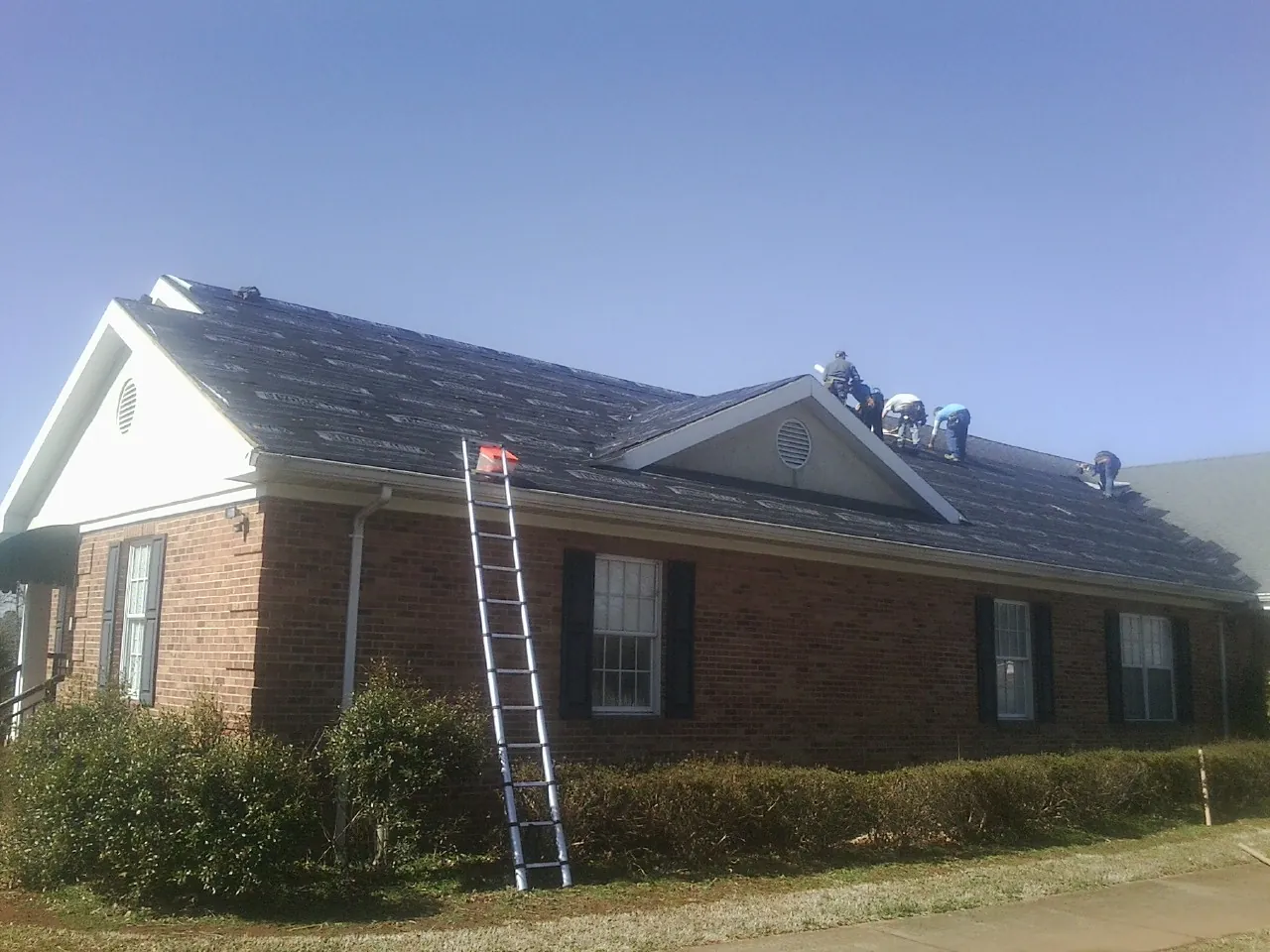 Crew tearing shingles off a brick ranch roof with a ladder set — Metal Roof Repair work by our crew in Whitt