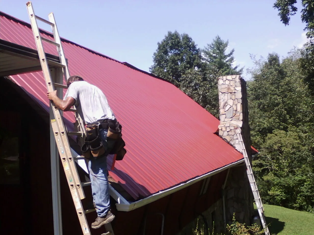 Roofer on a ladder beside a red metal roof and stone chimney — Soffit & Fascia Repair by Roofing Brothers in Whitt
